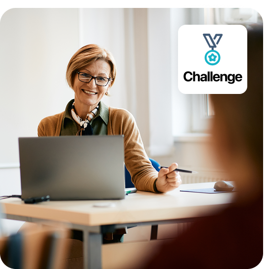 Image of teacher working behind a laptop in a classroom. She smiles at her students